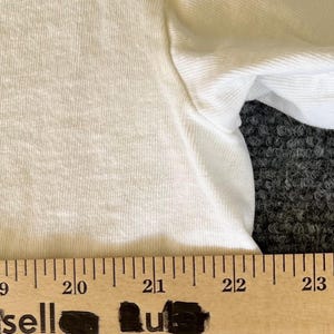 May include: Close-up of a white t-shirt with a visible texture. A wooden ruler with black markings is placed at the bottom of the shirt, with the word "sell" partially visible. The shirt's sleeve is also visible.
