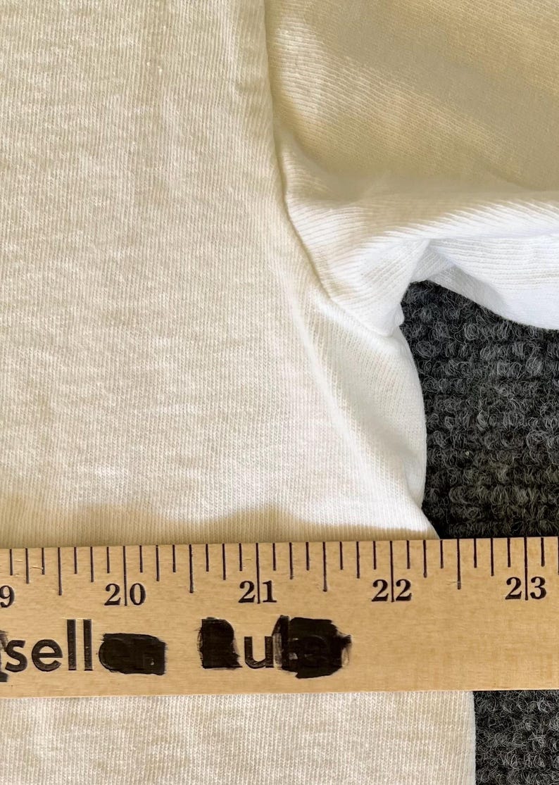 May include: Close-up of a white t-shirt with a visible texture. A wooden ruler with black markings is placed at the bottom of the shirt, with the word "sell" partially visible. The shirt's sleeve is also visible.