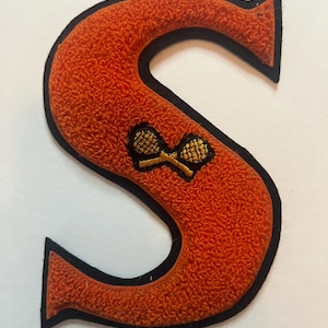 May include: A varsity-style letter S patch featuring a textured orange chenille surface with a black felt border. The center of the letter displays an embroidered graphic of two crossed gold tennis rackets with black netting details.