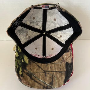May include: Camouflage baseball cap with a curved brim. The cap features a brown, green, and tan camouflage pattern on the exterior. The interior has a white lining with black stitching and a small American flag patch. A metal clasp adjusts the fit.
