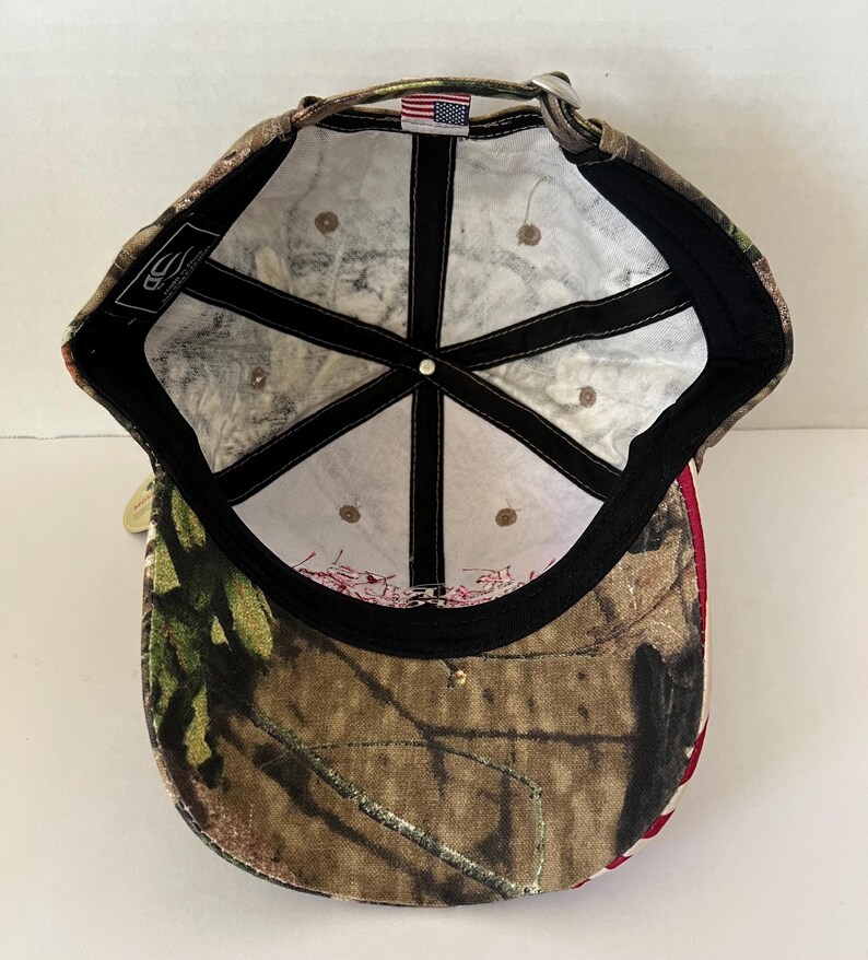 May include: Camouflage baseball cap with a curved brim. The cap features a brown, green, and tan camouflage pattern on the exterior. The interior has a white lining with black stitching and a small American flag patch. A metal clasp adjusts the fit.