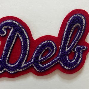 May include: A fabric patch featuring the word Del in purple cursive script with a white stitched outline, set against a red felt background. The lettering has a textured, raised appearance, and the entire piece is shaped to follow the contour of the text.