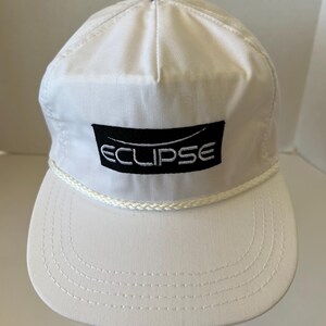 May include: White baseball cap with a curved brim and a rope detail. The cap features the word "ECLIPSE" in white lettering on a black rectangular patch. The cap is made of a lightweight material.