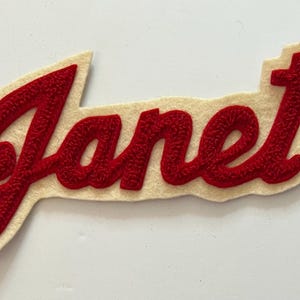 May include: A fabric patch featuring the name Janet written in a red cursive script with a textured, raised embroidery effect. The lettering is outlined by a cream-colored felt backing that follows the contour of the script, creating a border.
