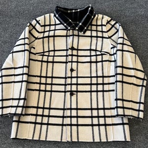 May include: A long-sleeved button-down jacket featuring a cream-colored base with a bold black grid pattern. The garment includes a structured collar, a front button closure with three visible dark buttons, and long sleeves.
