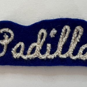 May include: A dark blue felt patch featuring the name Padilla embroidered in white thread. The lettering is in a cursive script style, with the white stitching standing out against the solid blue background of the fabric patch.