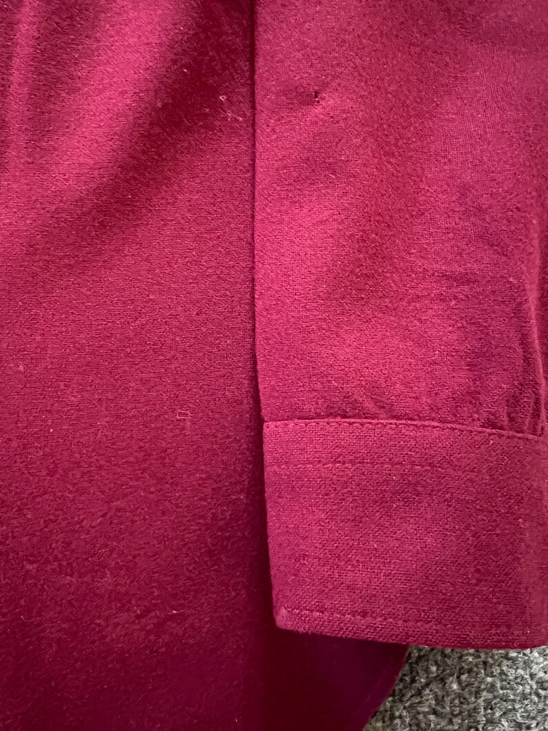 May include: A solid deep magenta long-sleeve shirt featuring a structured cuff with visible stitching. The fabric has a soft, matte texture and a consistent, uniform color throughout the sleeve and the cuff area, showcasing a clean design.