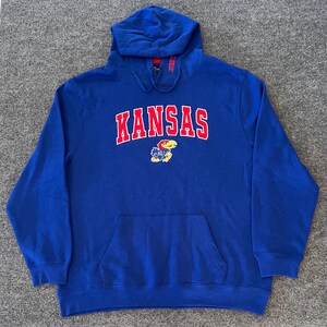 May include: A royal blue hooded sweatshirt with the word "KANSAS" in red block letters outlined in white across the chest. Below the word "KANSAS" is a small emblem. The sweatshirt has a kangaroo pocket and a drawstring hood.