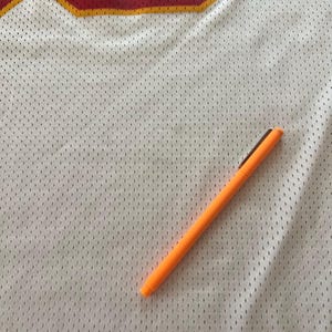 Op de afbeelding: Wit mesh sportshirt met rode en gouden accenten. Een oranje pen ligt diagonaal over het shirt. Het shirt heeft een groot nummer in de linkerbovenhoek.