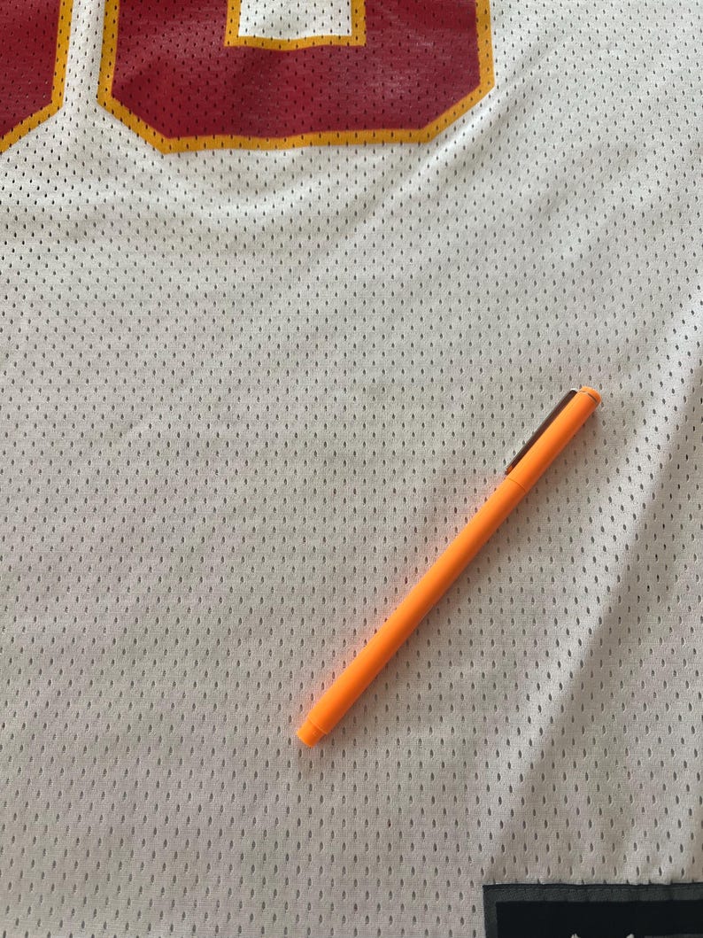 Op de afbeelding: Wit mesh sportshirt met rode en gouden accenten. Een oranje pen ligt diagonaal over het shirt. Het shirt heeft een groot nummer in de linkerbovenhoek.