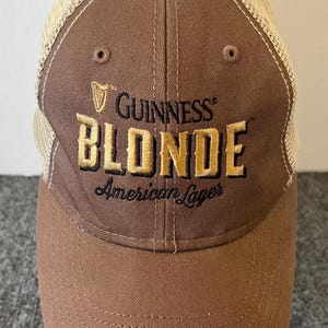 Op de afbeelding: Bruine baseballcap met een beige mesh achterkant. De voorkant van de cap heeft het Guinness-logo en de woorden "Guinness Blonde American Lager" geborduurd in goud en zwart.