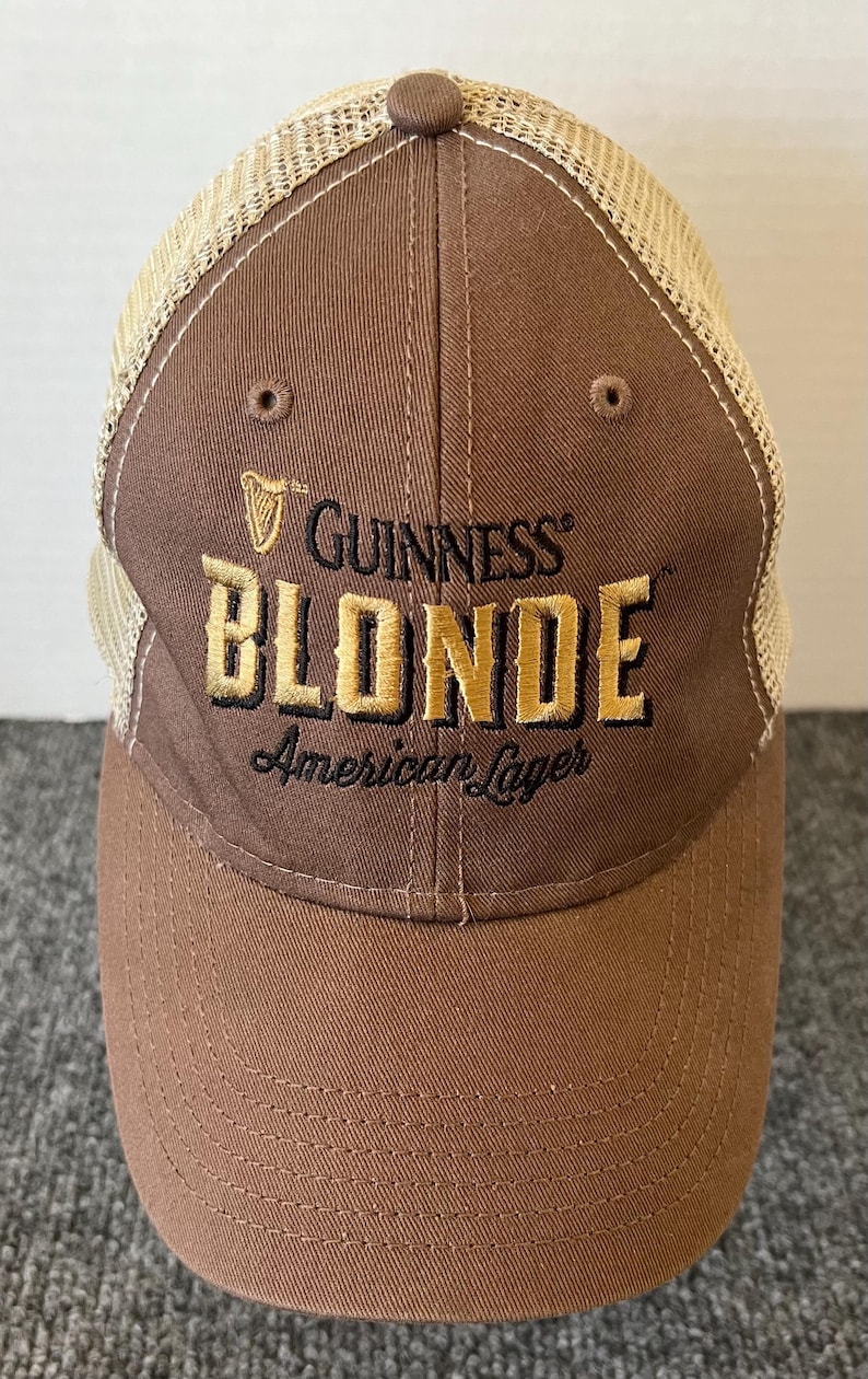 Op de afbeelding: Bruine baseballcap met een beige mesh achterkant. De voorkant van de cap heeft het Guinness-logo en de woorden "Guinness Blonde American Lager" geborduurd in goud en zwart.