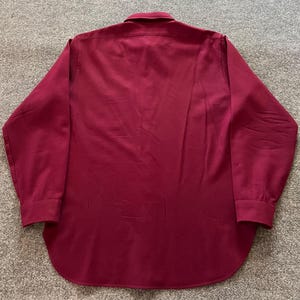 May include: A long-sleeve button-down shirt in a solid deep burgundy color. The shirt features a standard collar, a full-length button placket down the center, and buttoned cuffs at the end of each sleeve. The hem is rounded at the bottom.