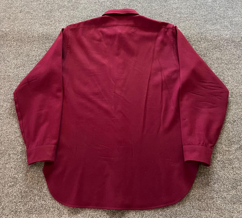 May include: A long-sleeve button-down shirt in a solid deep burgundy color. The shirt features a standard collar, a full-length button placket down the center, and buttoned cuffs at the end of each sleeve. The hem is rounded at the bottom.