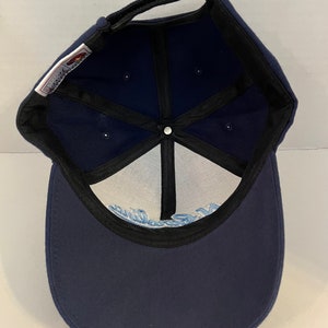 Pu&ograve; includere: Un cappello da baseball blu navy con visiera nera e un logo bianco all'interno. Il logo &egrave; un design a onda stilizzato con la parola "California" scritta in un font corsivo.