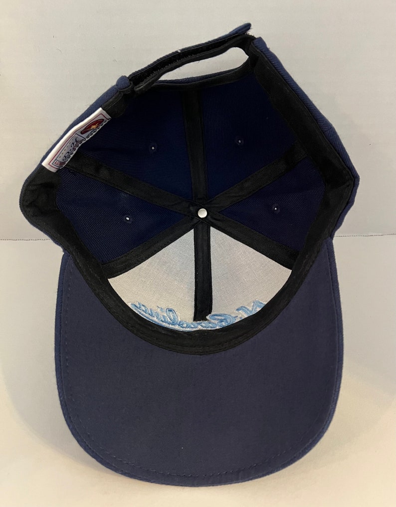 Pu&ograve; includere: Un cappello da baseball blu navy con visiera nera e un logo bianco all'interno. Il logo &egrave; un design a onda stilizzato con la parola "California" scritta in un font corsivo.