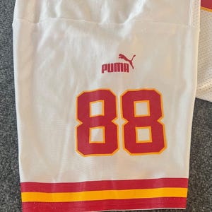 Op de afbeelding: Wit voetbalshirt met het nummer 88 in rood en geel. Het shirt heeft een rode en gele streep aan de onderkant en het Puma-logo in rood. Het shirt is gemaakt van een ademende, getextureerde stof.