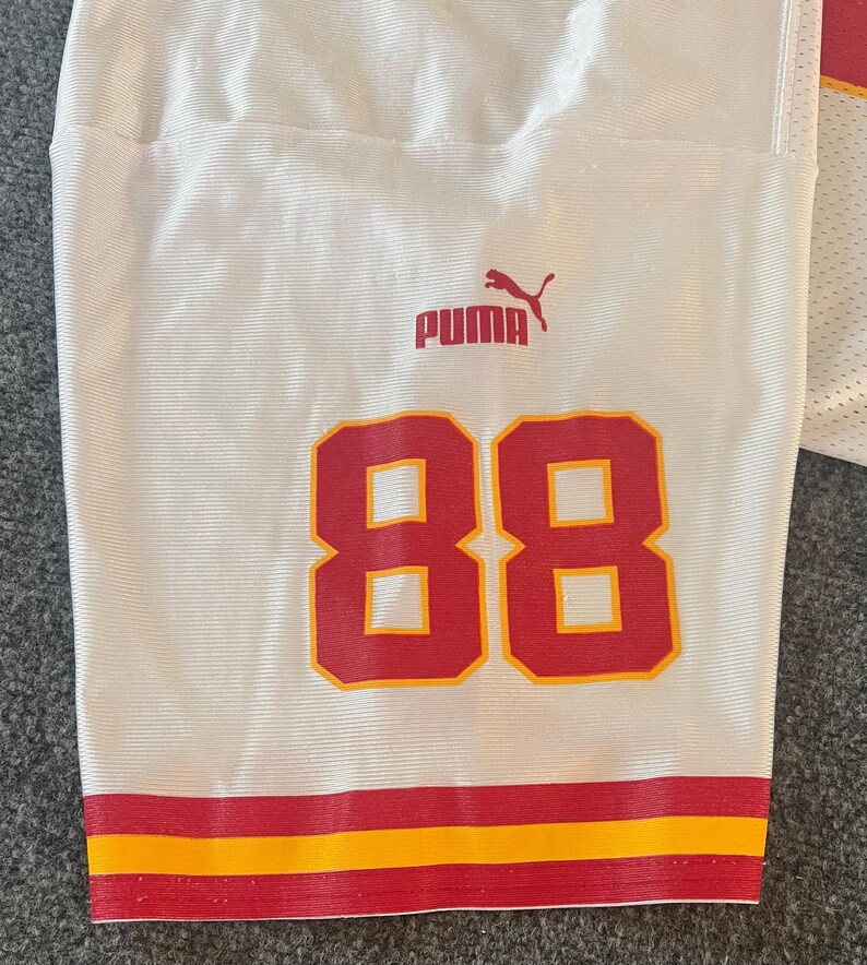 Op de afbeelding: Wit voetbalshirt met het nummer 88 in rood en geel. Het shirt heeft een rode en gele streep aan de onderkant en het Puma-logo in rood. Het shirt is gemaakt van een ademende, getextureerde stof.
