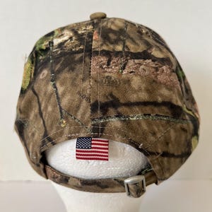 May include: Camouflage baseball cap with a curved brim. The cap features a brown, green, and tan pattern. An American flag tag is sewn on the back, and the adjustable strap has a silver buckle.