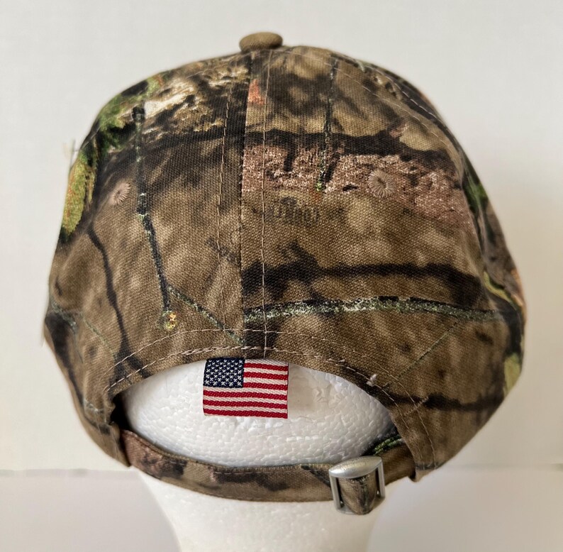 May include: Camouflage baseball cap with a curved brim. The cap features a brown, green, and tan pattern. An American flag tag is sewn on the back, and the adjustable strap has a silver buckle.