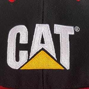 May include: Black baseball cap with a red brim and a white embroidered CAT logo with a yellow triangle.