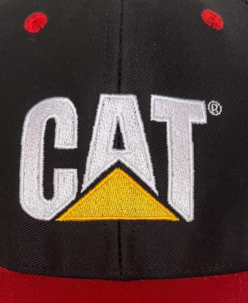 May include: Black baseball cap with a red brim and a white embroidered CAT logo with a yellow triangle.