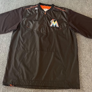 May include: Black short-sleeve baseball jacket with a half-zip closure. The jacket features the Miami Marlins logo on the chest and orange accents. The interior lining is orange.