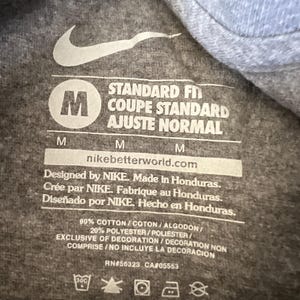Pu&ograve; includere: Primo piano di un'etichetta di una maglietta grigia con il logo Nike e testo. L'etichetta include la taglia "M", le istruzioni di lavaggio e il testo "VESTIBILIT&Agrave; STANDARD". La maglietta &egrave; in cotone e poliestere.