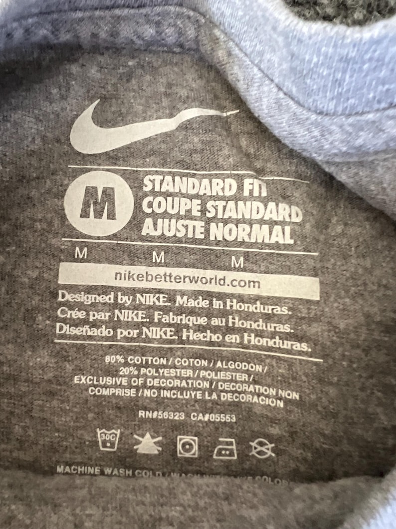 Pu&ograve; includere: Primo piano di un'etichetta di una maglietta grigia con il logo Nike e testo. L'etichetta include la taglia "M", le istruzioni di lavaggio e il testo "VESTIBILIT&Agrave; STANDARD". La maglietta &egrave; in cotone e poliestere.