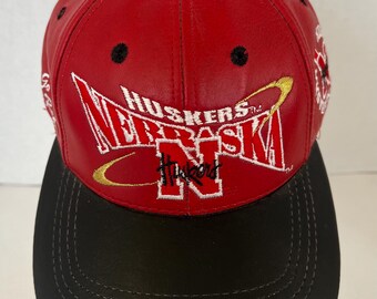 May include: A red and black baseball cap featuring the word "Huskers" in white and gold embroidery. The cap has a black brim and a black button on top. The cap is made of a leather-like material.