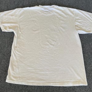 May include: A plain, off-white, short-sleeved t-shirt. The shirt is made of a soft, slightly wrinkled fabric. The t-shirt is laid flat on a gray surface.