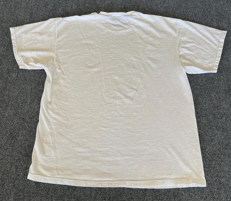May include: A plain, off-white, short-sleeved t-shirt. The shirt is made of a soft, slightly wrinkled fabric. The t-shirt is laid flat on a gray surface.