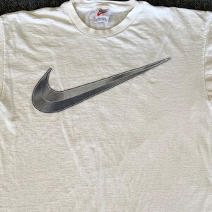 May include: Cream-colored t-shirt featuring a large, gradient gray Nike swoosh logo. The shirt has a crew neck and a small Nike tag near the collar. The fabric appears to be slightly textured.