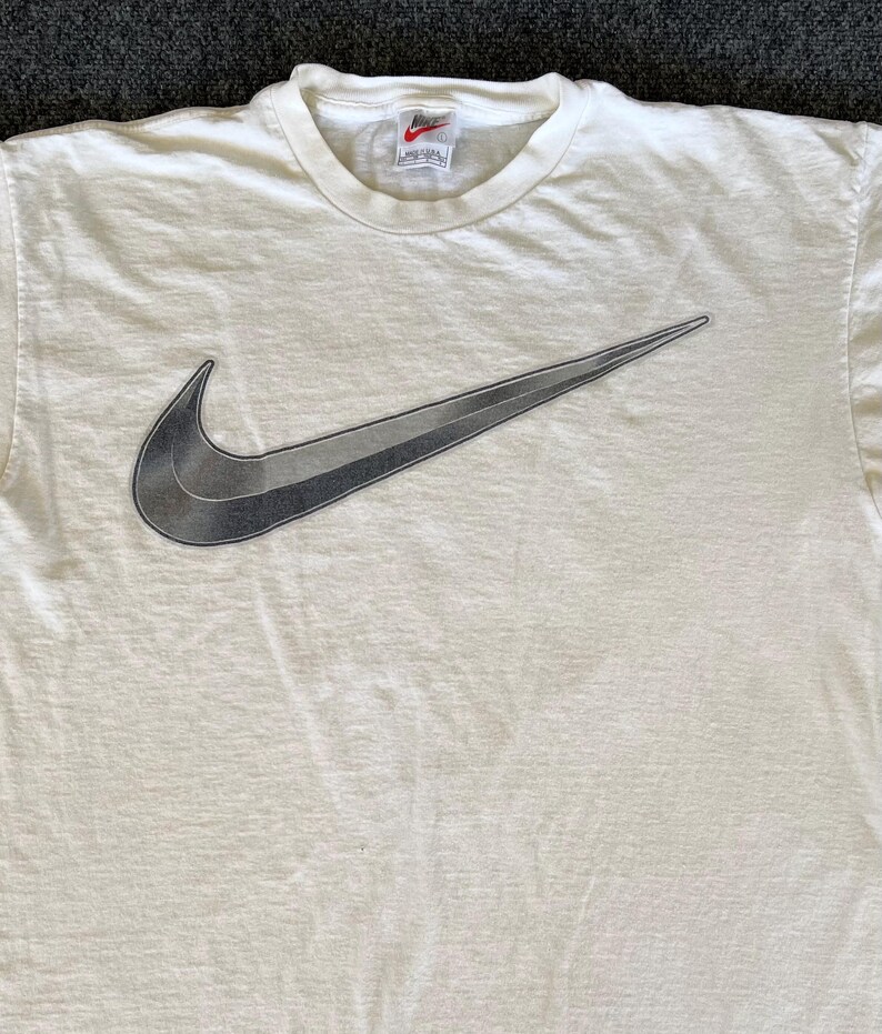 May include: Cream-colored t-shirt featuring a large, gradient gray Nike swoosh logo. The shirt has a crew neck and a small Nike tag near the collar. The fabric appears to be slightly textured.