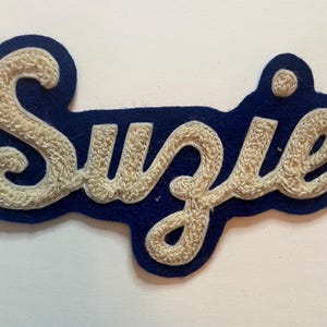 May include: A navy blue felt patch featuring the name Suzie embroidered in a cream-colored, textured chain stitch script font. The patch has a die-cut shape that follows the contour of the lettering, creating a bold and clear visual display.