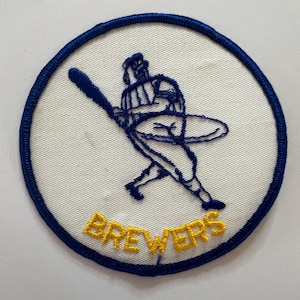 May include: A circular embroidered patch featuring a white background with a dark blue border. The center displays a dark blue stitched baseball player silhouette swinging a bat. Below the figure, the word BREWERS is stitched in yellow thread.