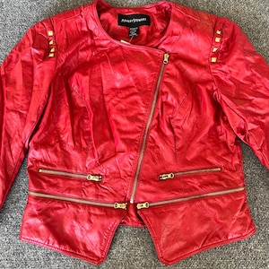 May include: A red Ashley Stewart jacket with a diagonal zipper and gold-colored zippers on the pockets. The jacket has gold studs on the shoulders and a quilted pattern on the lower front.