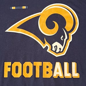 Op de afbeelding: Marineblauw t-shirt met een goud en wit ramshoofdlogo en het woord "FOOTBALL" in goud en witte blokletters. Een eenvoudig ontwerp, geschikt voor sportliefhebbers.