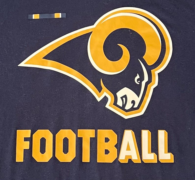 Op de afbeelding: Marineblauw t-shirt met een goud en wit ramshoofdlogo en het woord "FOOTBALL" in goud en witte blokletters. Een eenvoudig ontwerp, geschikt voor sportliefhebbers.