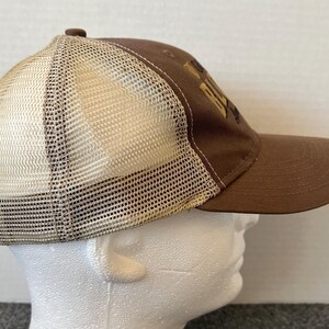 Op de afbeelding: Bruine truckerpet met een mesh achterkant en een gebogen klep. De pet heeft een bruin voorpaneel met goudkleurige geborduurde tekst. De mesh achterkant is lichtbruin en de pet heeft een bruine verstelbare band.