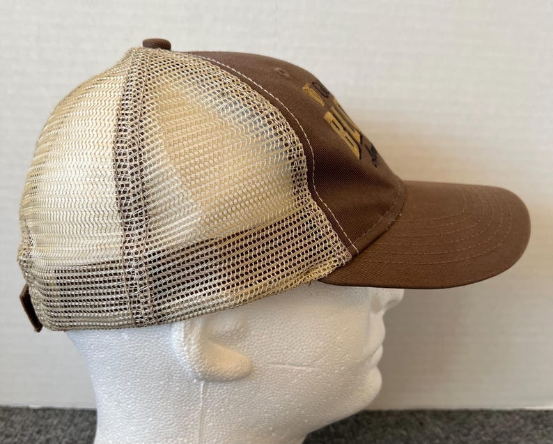 Op de afbeelding: Bruine truckerpet met een mesh achterkant en een gebogen klep. De pet heeft een bruin voorpaneel met goudkleurige geborduurde tekst. De mesh achterkant is lichtbruin en de pet heeft een bruine verstelbare band.