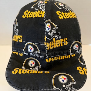 May include: A black cap featuring an all-over print of yellow and white Pittsburgh Steelers football helmet graphics and the word Steelers in yellow text. The cap has a central vertical seam with white zigzag stitching across the crown.