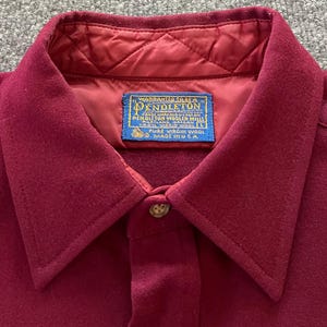 May include: A deep burgundy wool shirt featuring a quilted interior lining and a pointed collar. A blue rectangular label at the neck reads: Warranted to be a Pendleton, Pendleton Woolen Mills, Portland Oregon, 100% Virgin Wool, Made in USA.