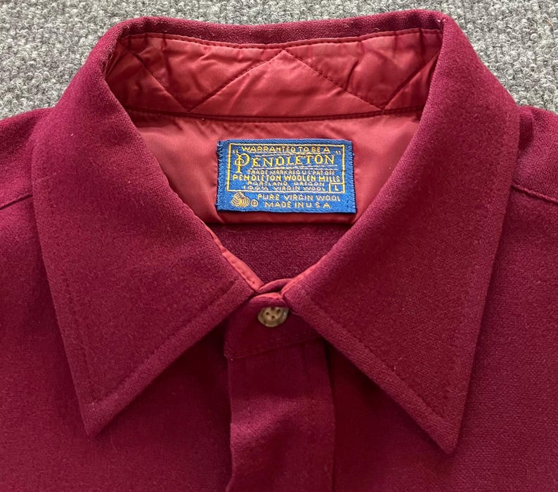 May include: A deep burgundy wool shirt featuring a quilted interior lining and a pointed collar. A blue rectangular label at the neck reads: Warranted to be a Pendleton, Pendleton Woolen Mills, Portland Oregon, 100% Virgin Wool, Made in USA.