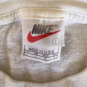 May include: Close-up of a white t-shirt collar with a Nike tag. The tag features the Nike logo in red and the text "MADE IN U.S.A." with size information. The shirt is a light gray color.
