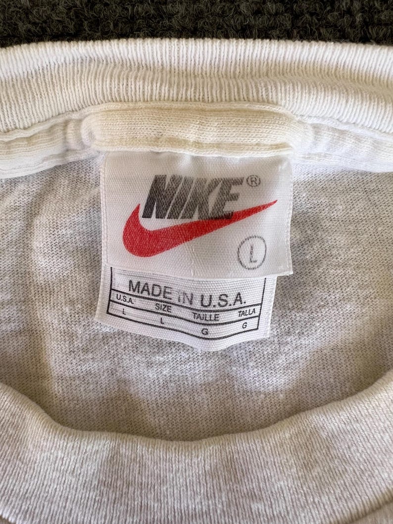 May include: Close-up of a white t-shirt collar with a Nike tag. The tag features the Nike logo in red and the text "MADE IN U.S.A." with size information. The shirt is a light gray color.