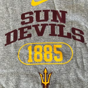 Pu&ograve; includere: T-shirt grigia con le parole "SUN DEVILS" in lettere maiuscole color marrone, con "1885" in un ovale dorato. Sono visibili anche un logo Nike dorato e un logo tridente dorato e marrone.