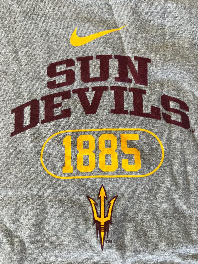 Pu&ograve; includere: T-shirt grigia con le parole "SUN DEVILS" in lettere maiuscole color marrone, con "1885" in un ovale dorato. Sono visibili anche un logo Nike dorato e un logo tridente dorato e marrone.