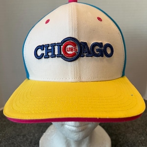 May include: A baseball cap with a white crown, a yellow brim, and a blue and pink accent. The word "CHICAGO" is embroidered in blue with a red and blue logo. The cap has a curved brim.