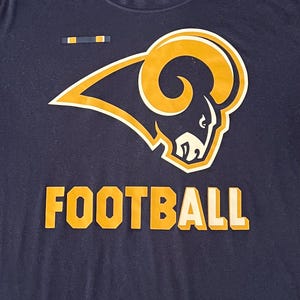 Op de afbeelding: Marineblauw T-shirt met het Los Angeles Rams logo in goud en wit, en het woord "FOOTBALL" in gouden blokletters. Het shirt heeft een ronde hals en korte mouwen. Het NFL-logo is zichtbaar.